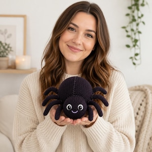 Puede incluir: Un peluche de araña tejido en color morado oscuro con ocho patas, un cuerpo redondo y dos ojos grandes blancos con pupilas negras. La araña presenta una pequeña boca bordada en color morado claro curvada en una sonrisa amigable.