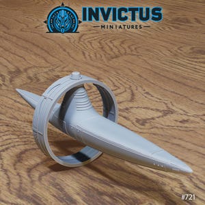 Op de afbeelding: Een lichtgrijs miniatuur ruimteschipmodel met een cirkelvormige ring en een puntige voorkant. Het model heeft de tekst "INVICTUS - MINIATURES" en het nummer "#721". Het ruimteschip staat op een houten oppervlak.