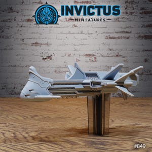 Op de afbeelding: Een lichtgrijs miniatuur ruimteschipmodel met een futuristisch ontwerp, met meerdere vleugels en een gedetailleerd lichaam. Het model wordt weergegeven op een heldere standaard tegen een bakstenen muurachtergrond. De tekst "INVICTUS MINIATURES" is zichtbaar bovenaan.