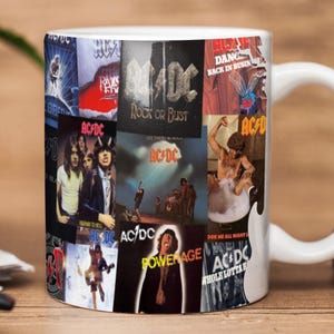 May include: White ceramic mug with a collage of AC/DC album covers. The mug displays album art such as "Rock or Bust" and "Highway to Hell." The handle is white, and a guitar shape is visible on the side.