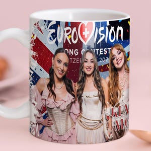Pode incluir: Caneca de cerâmica branca com o logotipo da Eurovisão e três mulheres em trajes de noite. O design incorpora uma bandeira da Union Jack. A caneca tem uma alça curva e está sobre um fundo rosa.