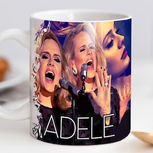 May include: White ceramic mug featuring multiple images of Adele singing, with the name "ADELE" in white lettering. The mug showcases the singer in a black dress, with a microphone, and a decorative design on the side.