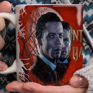 May include: White ceramic mug with a portrait of two men with glowing eyes set against a red background with bare tree branches. The mug features the word "INTUA" in gold lettering. The theme suggests fantasy or horror.