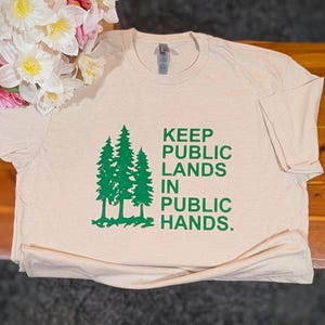 May include: A beige t-shirt with the text "KEEP PUBLIC LANDS IN PUBLIC HANDS" in green. The shirt features a graphic of three green pine trees. The shirt is made of a soft material and is folded on a wooden surface.