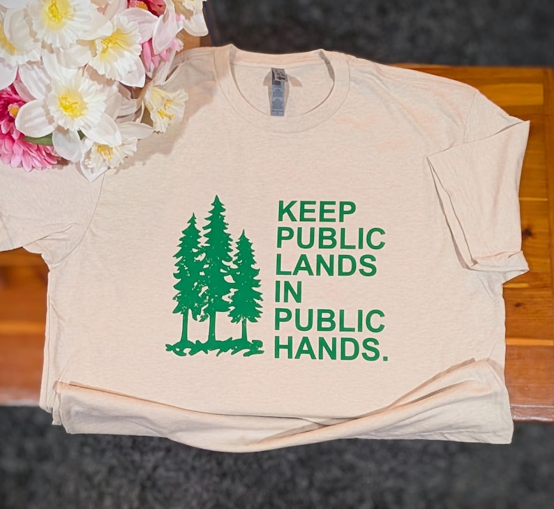 May include: A beige t-shirt with the text "KEEP PUBLIC LANDS IN PUBLIC HANDS" in green. The shirt features a graphic of three green pine trees. The shirt is made of a soft material and is folded on a wooden surface.