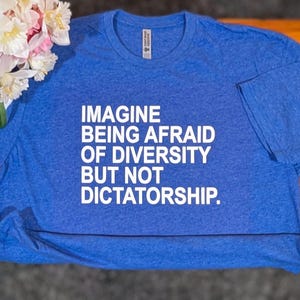 May include: A blue t-shirt with the text "IMAGINE BEING AFRAID OF DIVERSITY BUT NOT DICTATORSHIP" in white. The shirt is made of a soft material and is displayed on a wooden surface. A bouquet of white and pink flowers is in the upper left corner.