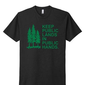 May include: Dark gray t-shirt with a green graphic of three pine trees and the text "KEEP PUBLIC LANDS IN PUBLIC HANDS." The shirt has short sleeves and a crew neck.