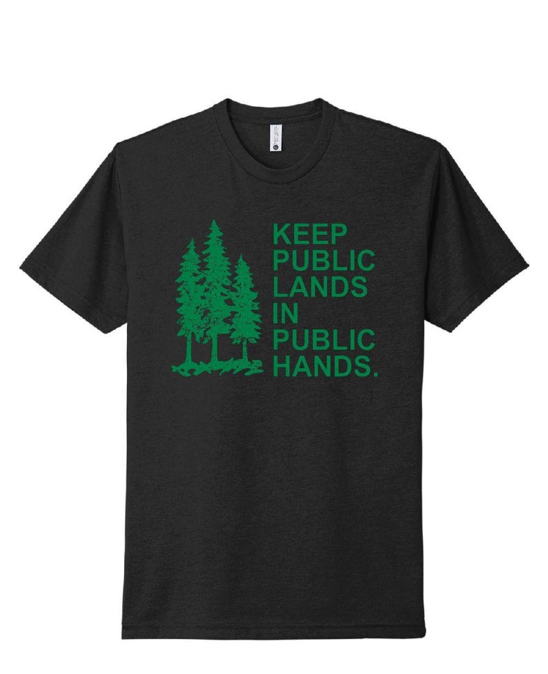 May include: Dark gray t-shirt with a green graphic of three pine trees and the text "KEEP PUBLIC LANDS IN PUBLIC HANDS." The shirt has short sleeves and a crew neck.
