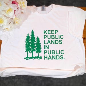 May include: White t-shirt with a green graphic of three pine trees and the text "KEEP PUBLIC LANDS IN PUBLIC HANDS." The shirt is made of a soft material and has a crew neck.