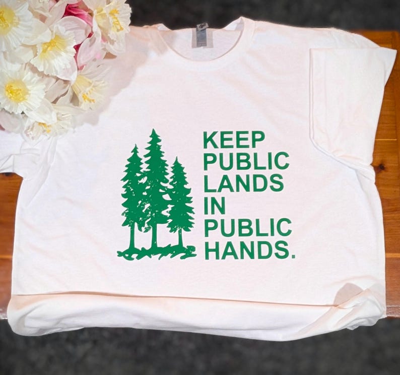 May include: White t-shirt with a green graphic of three pine trees and the text "KEEP PUBLIC LANDS IN PUBLIC HANDS." The shirt is made of a soft material and has a crew neck.