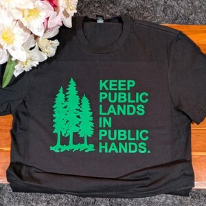 May include: Black t-shirt with green text and a tree graphic. The text reads "KEEP PUBLIC LANDS IN PUBLIC HANDS." The shirt is made of a soft material and is suitable for casual wear. A bouquet of flowers is in the upper left corner.