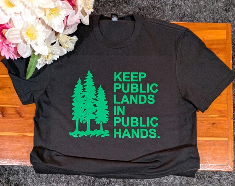 May include: Black t-shirt with green text and a tree graphic. The text reads "KEEP PUBLIC LANDS IN PUBLIC HANDS." The shirt is made of a soft material and is suitable for casual wear. A bouquet of flowers is in the upper left corner.