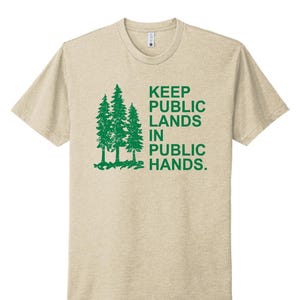 May include: Beige t-shirt with a green graphic of three pine trees and the text "KEEP PUBLIC LANDS IN PUBLIC HANDS." The shirt has a crew neck and short sleeves. The design is centered on the front of the shirt.