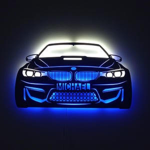 Peut inclure: Décoration murale en forme de voiture noire avec des lumières LED bleues. L'avant de la voiture présente des phares, une calandre et le nom "MICHAEL" illuminé en bleu. L'œuvre est conçue pour être accrochée au mur et a un effet lumineux.