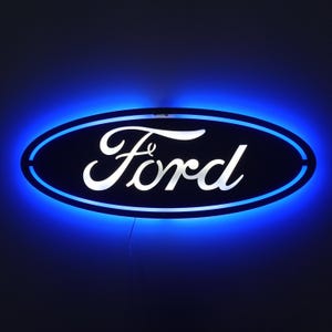 May include: A Ford logo sign illuminated with blue LED lights. The sign features the Ford oval in dark blue with the word "Ford" in white. The sign is mounted on a dark surface, creating a striking contrast.
