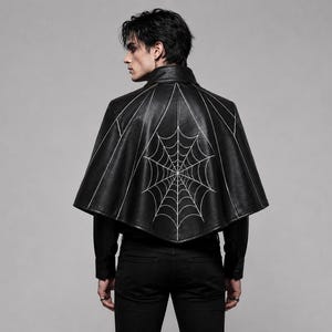 May include: A black capelet featuring a white embroidered spiderweb design on the back. The garment has a high collar, a flared silhouette, and is worn over a black long-sleeved shirt and black trousers. The design includes radiating lines.