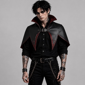 May include: A black capelet featuring a high collar with a deep red interior lining, secured at the front with a buckle strap. The garment is worn over a black button-down shirt, paired with black trousers featuring metal chains and a leather belt.