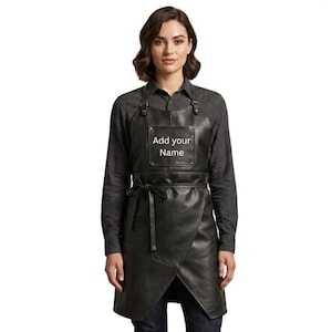 May include: A black leather apron with a wrap-style design. The apron has adjustable straps and a tie at the waist. The text "Add your Name" is displayed on the chest area. The model is wearing a dark gray long-sleeve shirt and dark blue jeans.