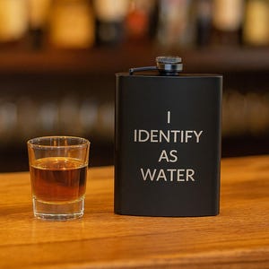 Może przedstawiać: Czarny flakon z napisem "I IDENTIFY AS WATER" w srebrnych literach, obok kieliszka wypełnionego bursztynowym płynem. Flakon ma małą, czarną zakrętkę i stoi na drewnianej powierzchni.