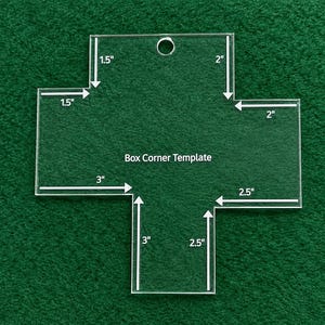 May include: A clear acrylic cross-shaped box corner template with a hole at the top. The template features white printed text reading Box Corner Template and white arrows indicating measurements of 1.5 inches, 2 inches, 3 inches, and 2.5 inches.