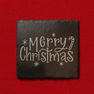 Könnte beinhalten: Ein schwarzer quadratischer Schieferuntersetzer mit dem Schriftzug "Merry Christmas" in einer dekorativen Schriftart. Der Text ist hell und von Sternen und einer Zuckerstange umgeben. Der Untersetzer befindet sich auf einem roten, strukturierten Hintergrund.