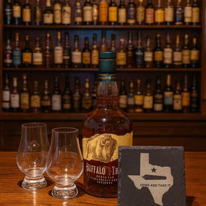 Könnte beinhalten: Eine Flasche Buffalo Trace Bourbon mit zwei Gläsern und einem Texas-förmigen Untersetzer mit der Aufschrift "Come and Take It". Die Bourbon-Flasche hat ein goldenes Etikett mit einem Bison-Bild. Im Hintergrund Regale mit Likörflaschen.
