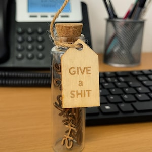 Könnte beinhalten: Eine klare Glasflasche mit Korken und einem Holzanhänger mit der Aufschrift "GIVE a SHIT". Die Flasche enthält kleine Holzausschnitte des Wortes "SHAPE". Der Anhänger ist mit Bindfaden an der Flasche befestigt.