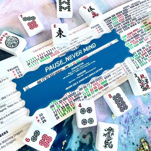 Könnte beinhalten: Weiße Mahjong-Steine mit roten und schwarzen Symbolen, darunter Zahlen, Zeichen und Drachen, sind auf einer gemusterten Oberfläche verstreut. Eine blaue Karte mit dem Text "PAUSE...NEVER MIND" ist ebenfalls vorhanden, zusammen mit einer Spielanleitung.