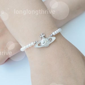 May include: A pearl bracelet with a silver-tone charm. The charm is a stylized orb design with a cross on top, encrusted with small, clear crystals. The bracelet is strung with small, round, white pearls. The text "longlongthrive" is in the background.