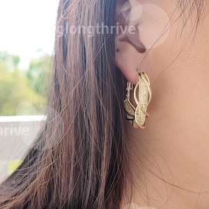 May include: Gold-toned hoop earrings with a woven design. The earrings feature a textured, open weave pattern and are worn on a person's ear. The earrings are a jewelry item.