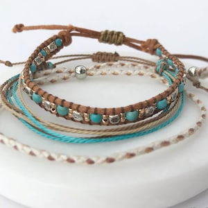 May include: A collection of handcrafted bracelets in different designs and colours. The bracelets feature brown leather, turquoise beads, and braided cords in shades of beige, white, and teal. The bracelets are presented on a white, circular surface.