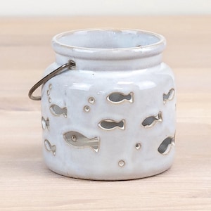 May include: A white ceramic candle holder with a glossy finish, featuring cut-out fish and bubble shapes. The jar has a rounded body, a wide opening, and a thin dark metal wire handle attached to the sides. The text Harbour Hut DESIGNS is visible.