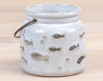 Puede incluir: Un portavelas de cerámica blanca con acabado brillante, que presenta recortes en forma de peces y burbujas. El tarro tiene un cuerpo redondeado, una abertura ancha y un asa de alambre de metal oscuro. El texto Harbour Hut DESIGNS es visible.