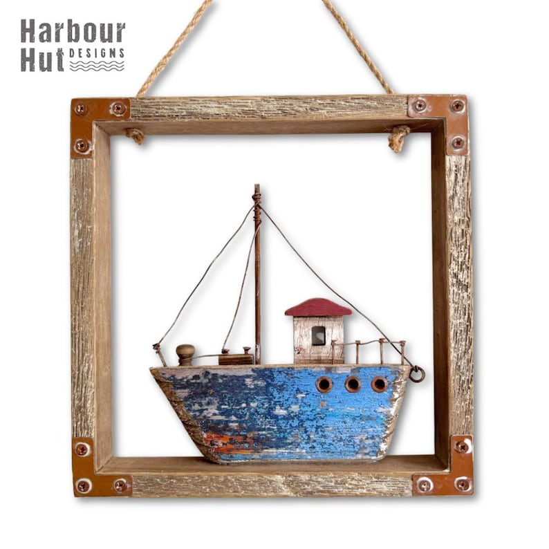May include: A square wooden frame with copper-colored corner brackets containing a model fishing boat. The boat has a weathered blue hull, a tall mast with thin wire rigging, a small red roof cabin, and three circular portholes on the side.