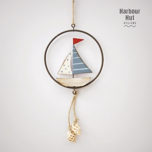 May include: A wooden sailboat with blue and white stripes and white polka dots hangs from a metal ring with a brown rope and two seashells. The sailboat has a red sail. The text "Harbour Hut Designs" is in the top right corner of the image.