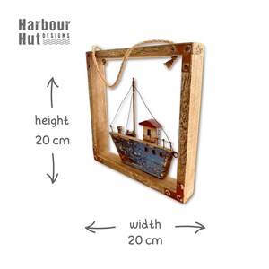 May include: A square wooden frame containing a model fishing boat with a blue hull and red cabin. The frame features metal corner accents and a jute hanging rope. Text indicates a height of 20 cm and a width of 20 cm. The logo Harbour Hut Designs is visible.