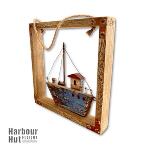 May include: A square wooden frame with a rustic finish and metal corner accents, featuring a hanging jute rope. Inside the frame is a model fishing boat with a weathered blue hull, a red roof, and thin metal masts with black rigging lines.