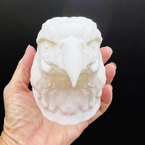 May include: White ceramic sculpture of an eagle's head. The sculpture is detailed and realistic, with a sharp beak and intense eyes.