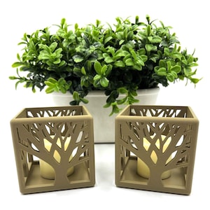 May include: Two brown candle holders with a tree design. The candle holders are made of a textured material and have a square shape. Each holder has a small, white candle inside.