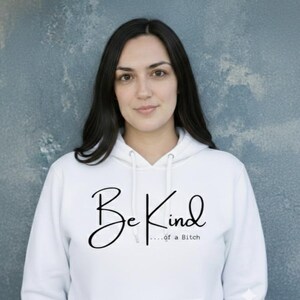 Puede incluir: Sudadera con capucha blanca con el texto "Be Kind...of a Bitch" en escritura negra. La sudadera tiene una capucha con cordón y un bolsillo delantero. El fondo es una pared azul-gris texturizada.