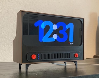 Peut inclure: Une horloge numérique de style rétro, avec un boîtier marron et noir rappelant un ancien téléviseur. L'écran affiche l'heure 12:31 en bleu. Elle possède des boutons rouges et de petits pieds noirs.