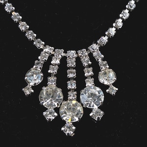 Op de afbeelding: Een zilverkleurige ketting met heldere, sprankelende strass-steentjes. De ketting heeft een centrale hanger met grotere, ronde stenen die naar beneden vallen, wat een glamoureus ontwerp creëert. De ketting staat tegen een zwarte achtergrond.