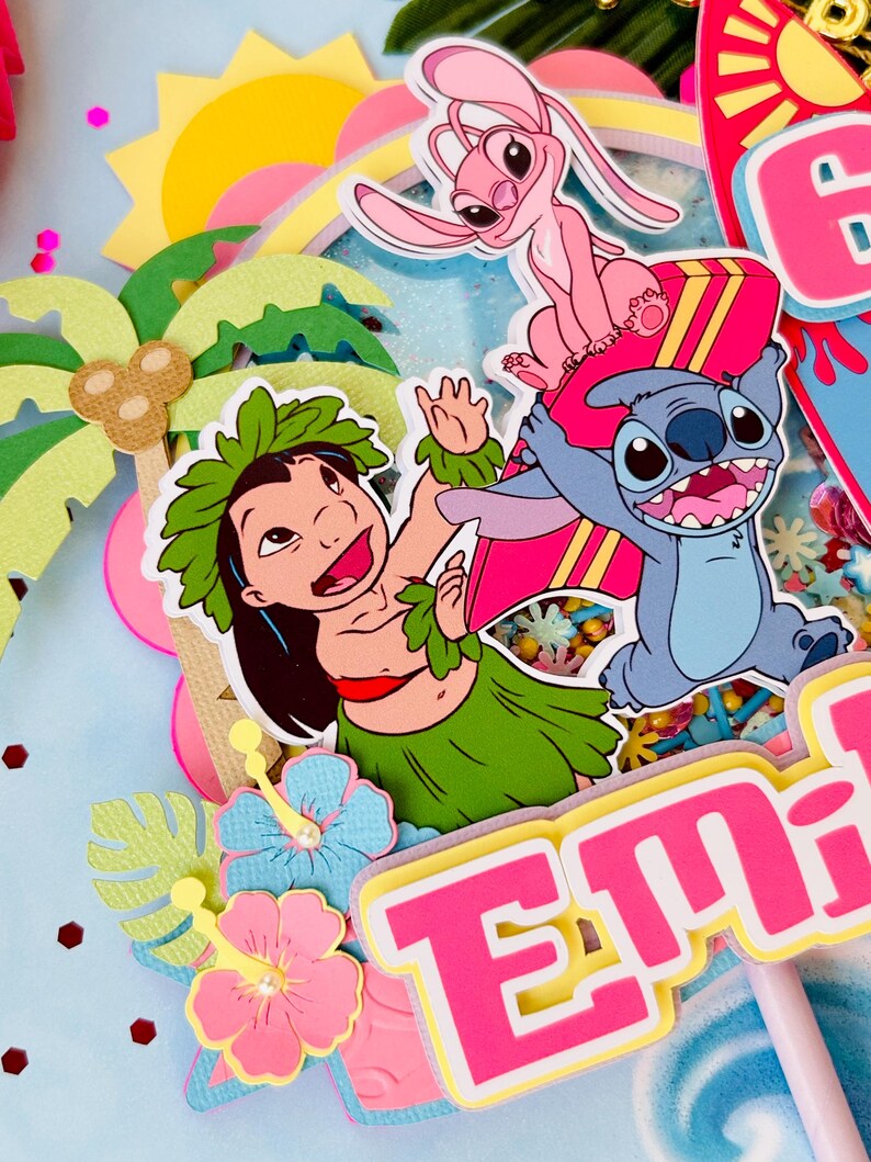 K&ouml;nnte beinhalten: Ein farbenfroher Tortenaufsatz mit den Zeichentrickfiguren Lilo, Stitch und Angel. Das Design umfasst einen rosa und gelben Namen, Palmen, Hibiskusbl&uuml;ten und eine Sonne. Der Aufsatz ist mit Glitzer und Konfetti verziert.