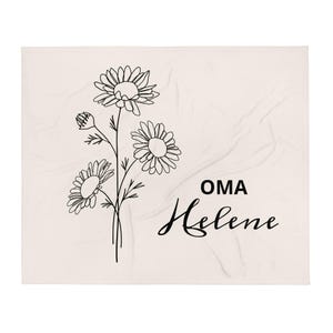 Könnte beinhalten: Eine cremefarbene Decke mit einer schwarzen Strichzeichnung von Gänseblümchen. Die Decke trägt die Worte "OMA Helene" in schwarzer Schrift. Die Gänseblümchen befinden sich in verschiedenen Blühstadien mit detaillierten Blütenblättern und Stielen.