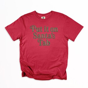 May include: A red t-shirt with the phrase "Put it on Santa's Tab" in green lettering. The shirt is made of a soft material and has a classic crew neck design. Perfect for the holiday season.