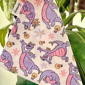 May include: White triangular fabric with a repeating pattern of a purple dragon, pink flowers, and light bulbs. The dragon has a pink belly and yellow accents. The fabric is held by a hand with purple nail polish. The tag says "Sew Trendy Co."
