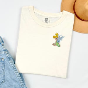 May include: Cream-colored t-shirt featuring a small embroidered image of a fairy with green and blue accents. The shirt is folded, with a tag visible at the neckline. A pair of blue denim overalls and a tan hat are partially visible in the background.