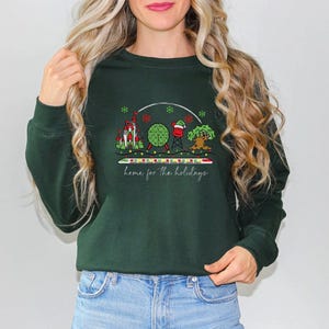 May include: A dark green sweatshirt with a colorful graphic of a castle, a globe, a red and white ball, and a tree, with the text "home for the holidays" in a script font. The sweatshirt is made of a soft material.