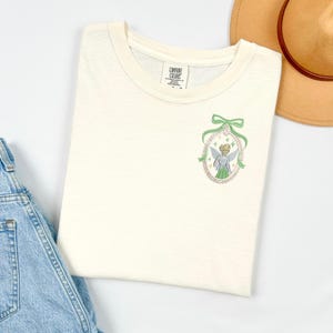 May include: Cream-coloured t-shirt with a round neckline. The shirt features an embroidered design of a fairy in a green and pink oval frame with a green bow. The shirt is made of a soft material and is folded on a white surface.