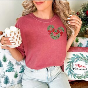 May include: A dusty rose-colored t-shirt featuring a Mickey Mouse-shaped wreath design with red bows. The person is holding a mug filled with marshmallows. The mug has a gingerbread man pattern. Christmas-themed pillows are in the background.